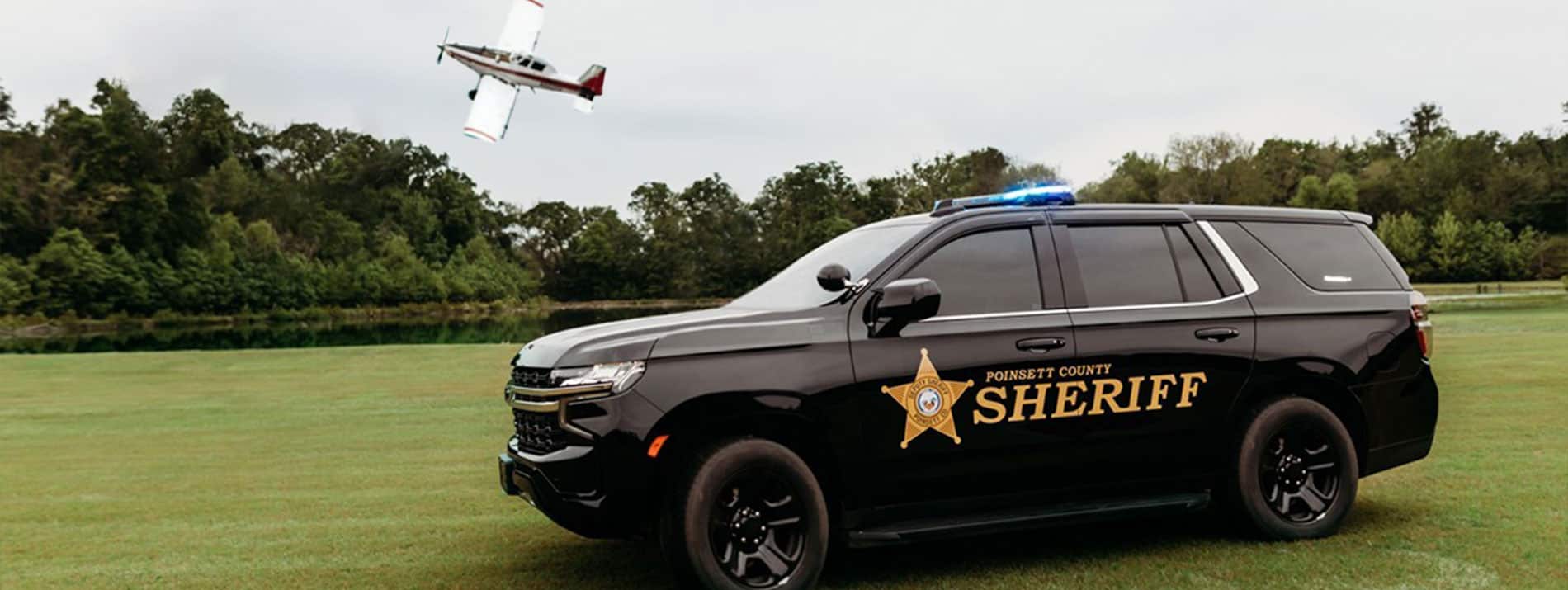 A Poinsett County Sheriff vehicle in a field with an airplane flying behind it.
